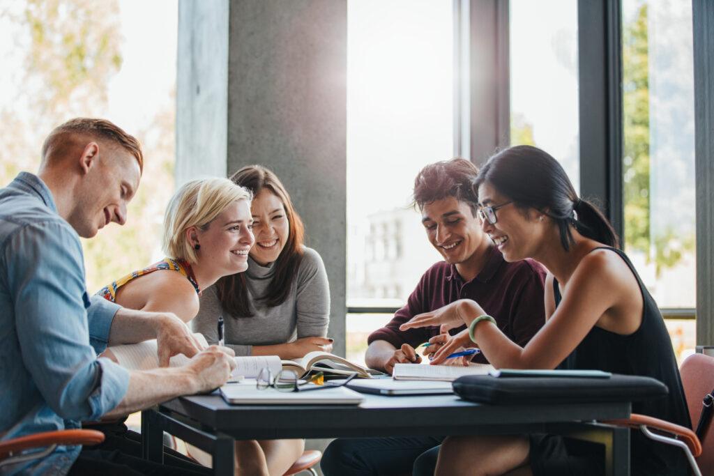 a group of young people sit around a table, holding pens, smiling and talking. A variety of notebooks and laptops are on the table, and a pair of glasses is placed on top of one of the notebooks.