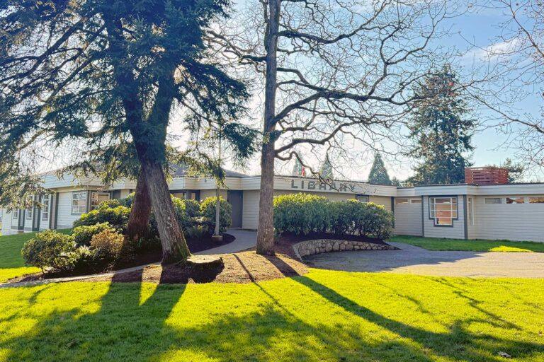 Exterior of the Central Saanich branch of the Greater Victoria Public Library on a sunny day, surrounded by mature trees and green lawns.