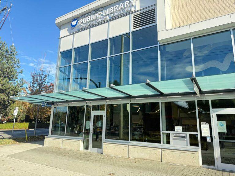 Exterior of the sxʷeŋxʷəŋ təŋəxʷ (James Bay) branch of the Greater Victoria Public Library, a modern glass-fronted building with a covered entrance canopy on a sunny day.