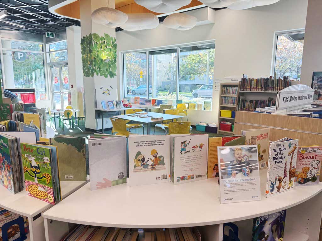 Cheery interior of the SJB branch - childrens books on a shelf display are seen in the foreground, and tables in a carpeted room are seen in the background. the library has large windows and lots of light