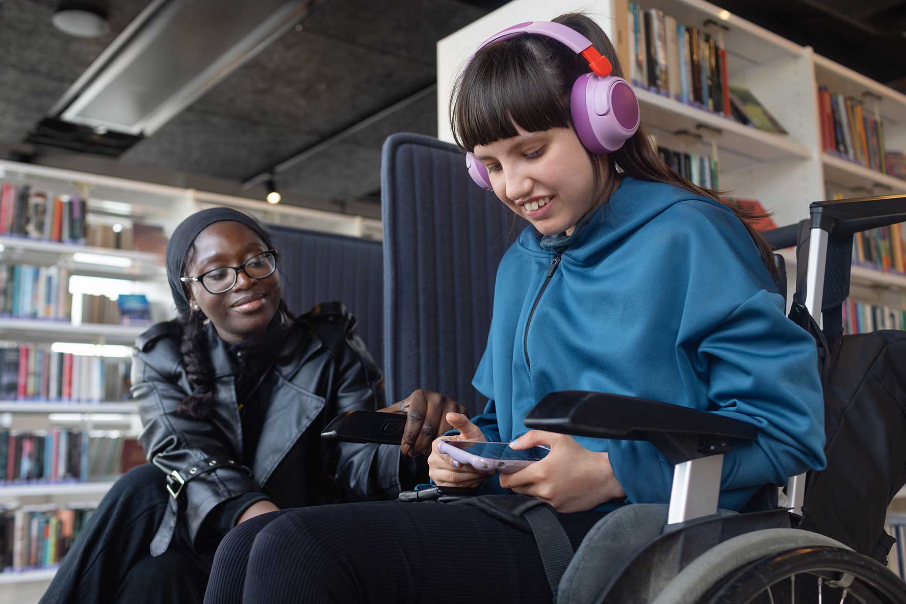 Smiling young girl in wheelchair with a tablet, a caregiver friend beside her smiling.