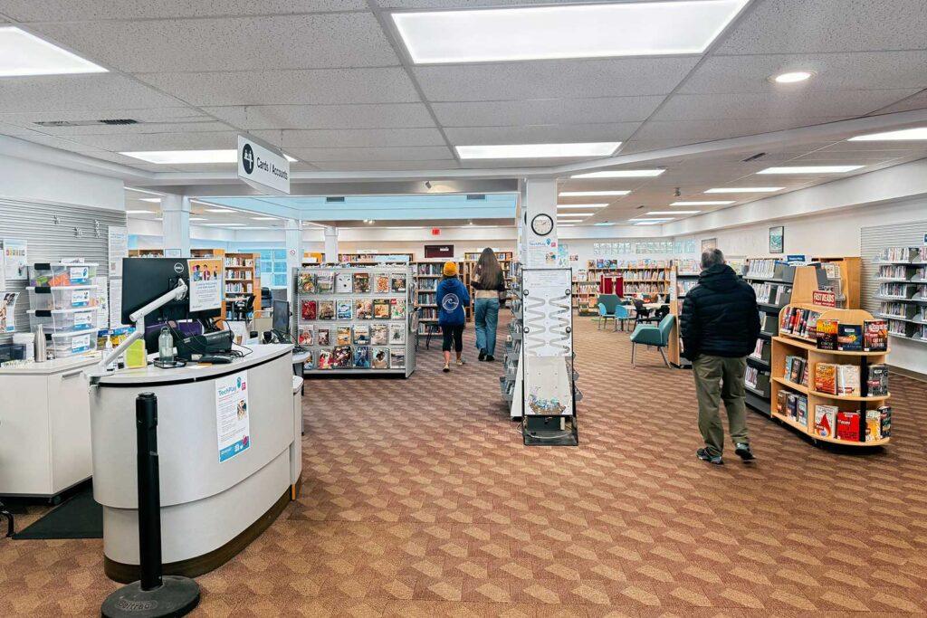 Interior of the Central Saanich branch showing the service desk, magazine displays, and book shelves, with library patrons browsing the collection.