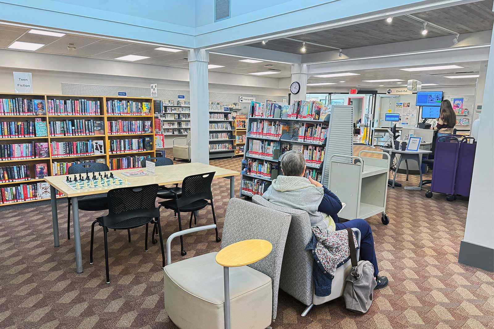Interior of the Central Saanich branch showing a seating area with comfortable chairs, a table with a chess set, and shelves of teen fiction books, with the checkout desk visible in the background.