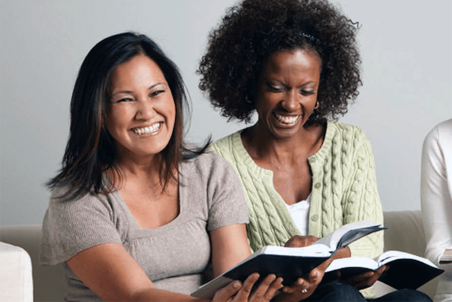 Two women sitting together laughing and reading from books.