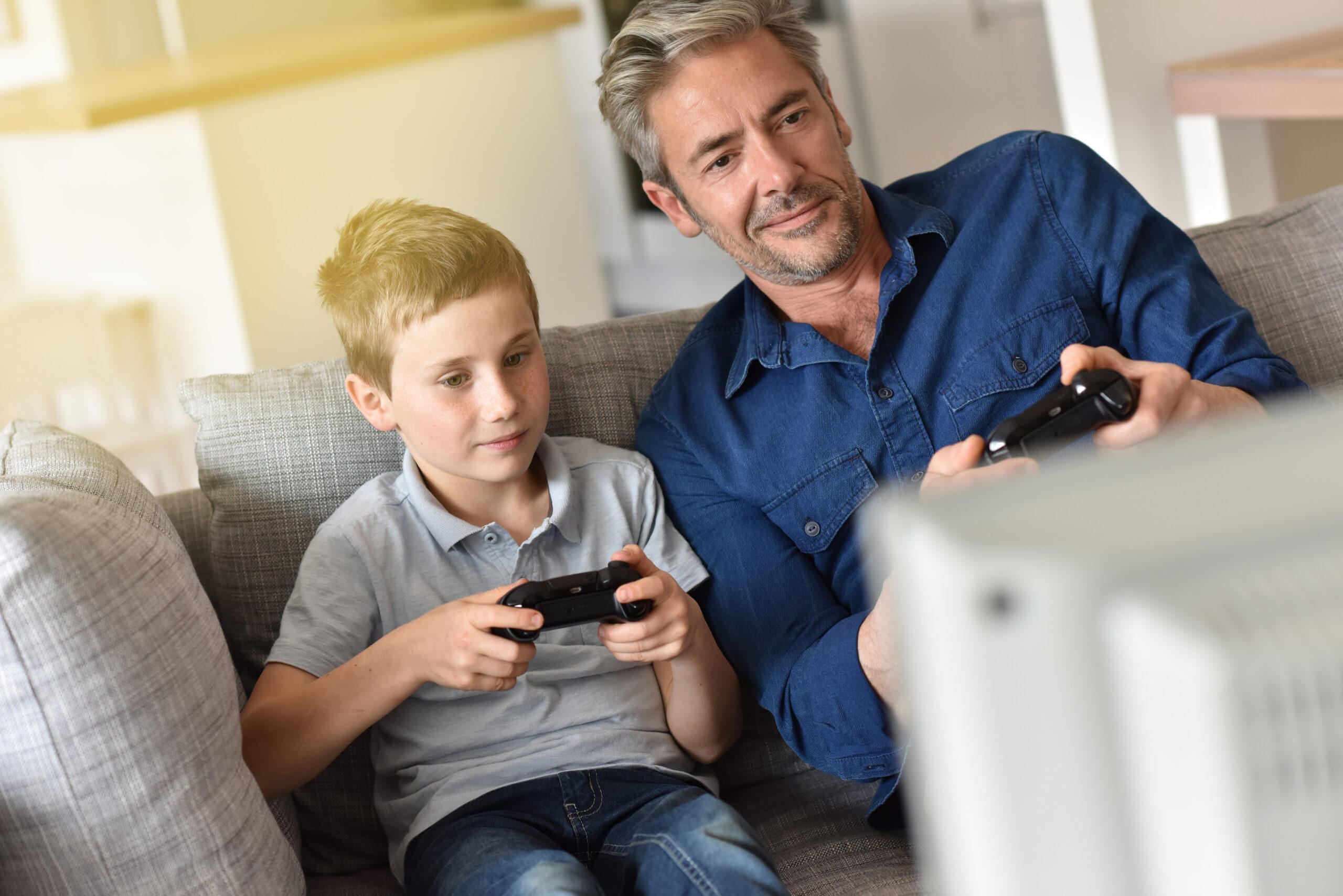 A man and a young boy sit together on a couch while holding game controllers and appear to be playing a video game.