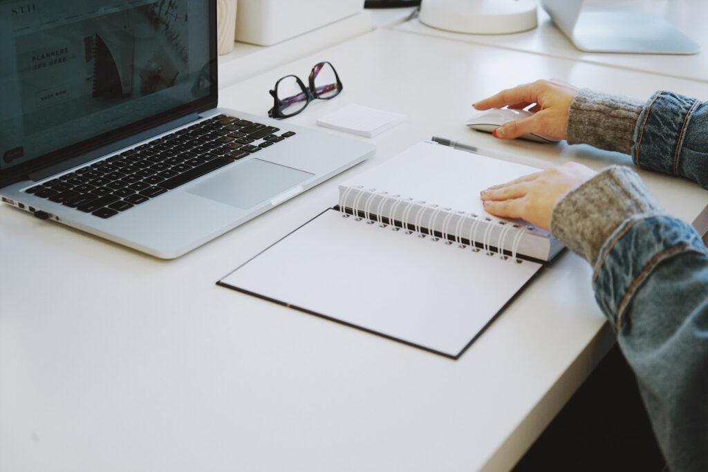 two hands turning a page in a notebook with a laptop and a pair of glasses on the  table