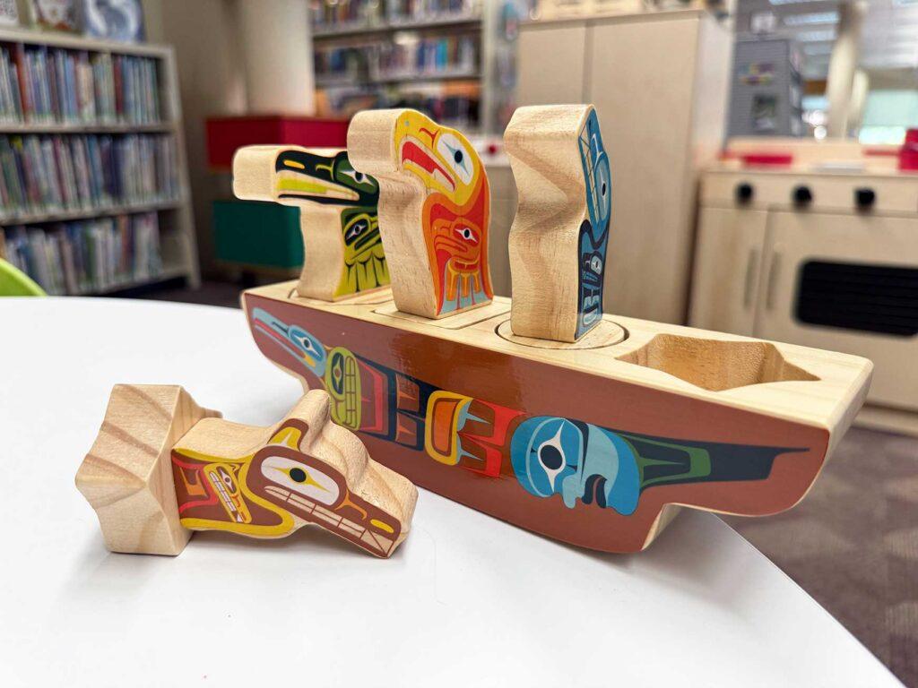 an indigenous wood toy puzzle. It is a shape of a wooden canoe with indigenous artworks painted on the sides, and the top of the canoe has different shaped holes into which wooden creatures can fit. The wooden pegs of the puzzle are shaped like animals with traditional designs painted on them