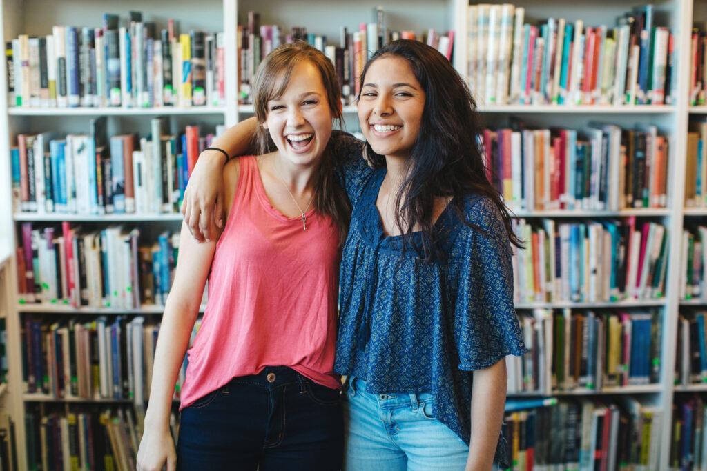 two young women stand in front of a library bookshelf. One has her arm around the others shoulder, and they are both smiling/laughing for the camera