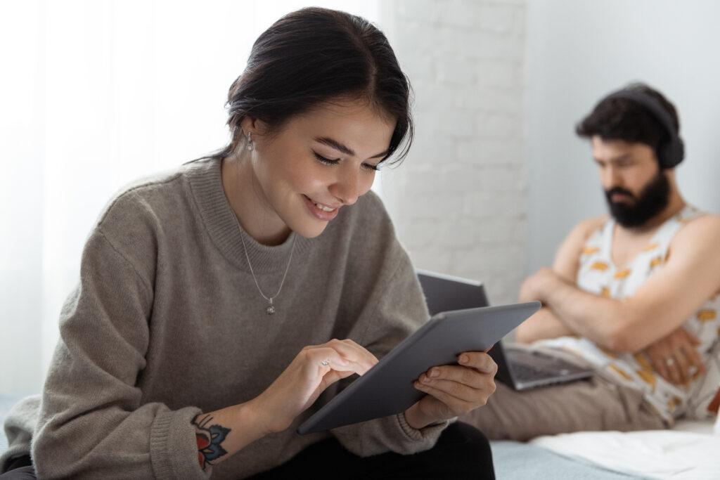 Young woman smiling and browsing tablet while resting on bed near man with laptop in weekend at home