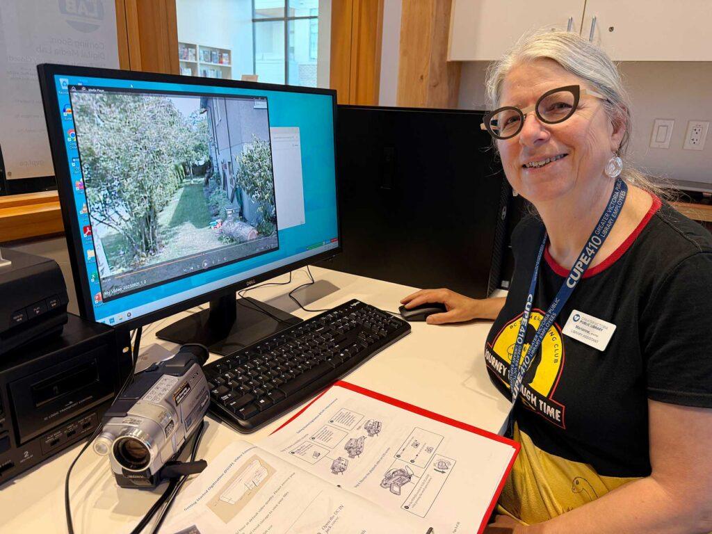 a library assistant sits at a computer and smiles at the camera. A video recorder hooked up to the computer is sitting on the desk in front of her and there is an instruction manual on the desk with images of the recorder. The computer monitor shows a media player window displaying an outdoor scene with a row of trees and the side of a building.