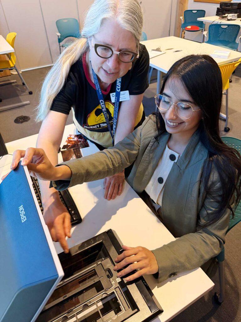 a library assistant is helping a young woman with some equipment. Epson logo is seen on the equipment and there are some old photographs and a keyboard seen in on the desk