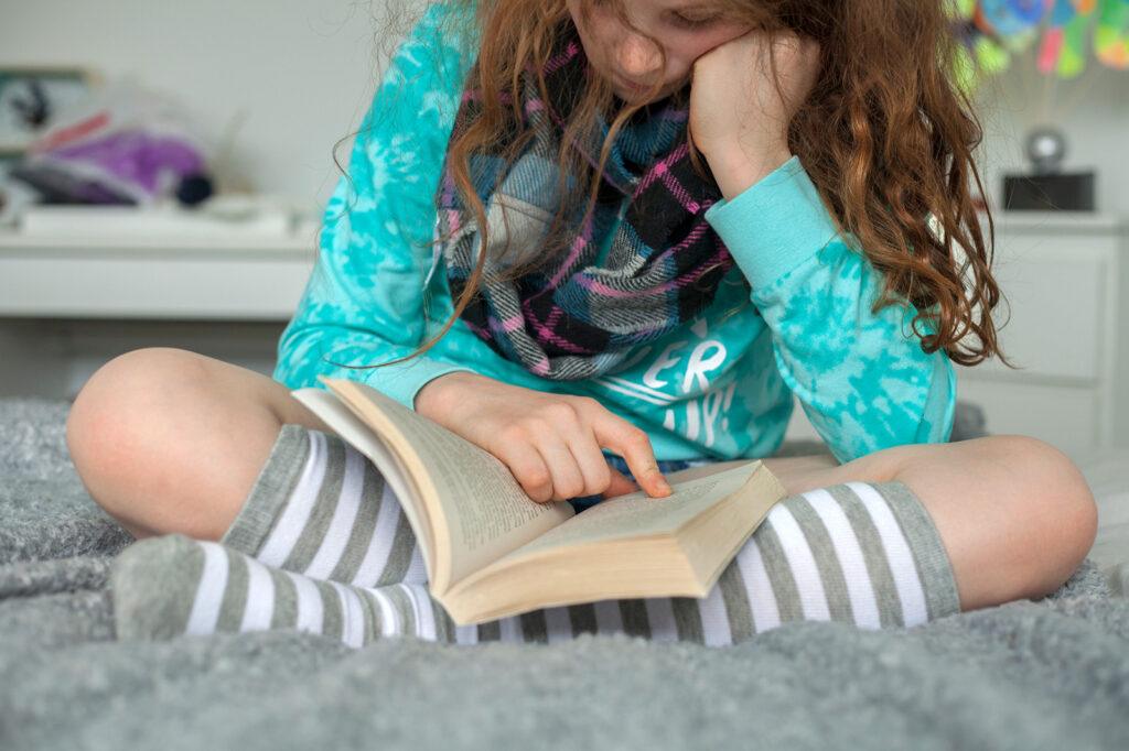 a young tween-aged girl is shown sitting cross-legged resting her face on her hand while reading a book