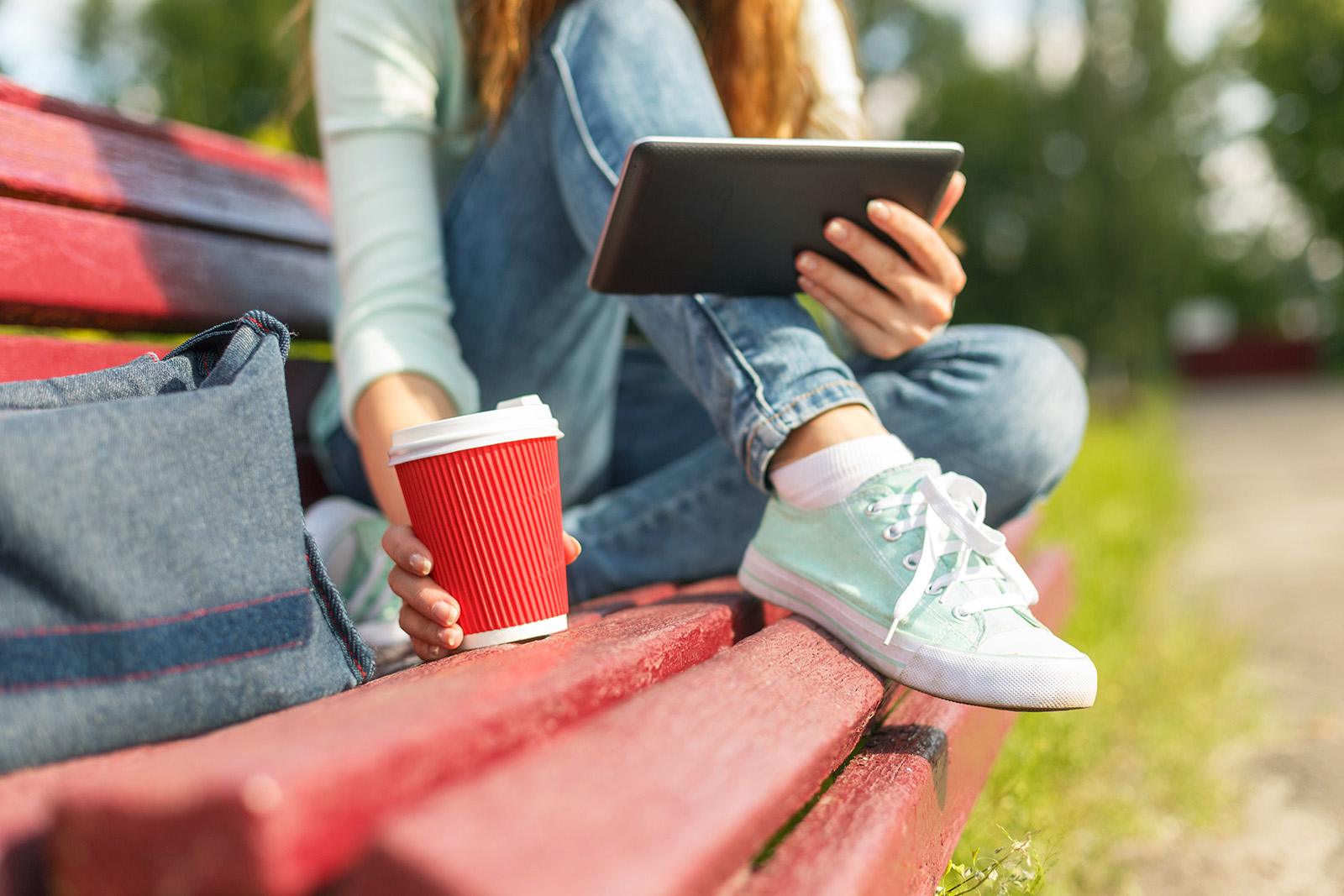 image of someone sitting on a park bench with a coffee cup reading on a tablet