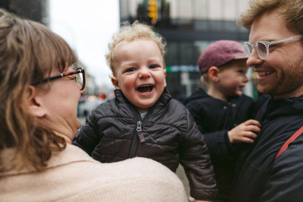 a smiling lady holding a laughing toddler next to a smiling man holding another toddler. It looks like they are outside on a street.