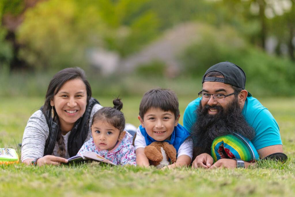 a young indigenous family is laying on a lawn. The mother is holding a book and two young children are in the middle. All of them are looking up from their reading and smiling at the camera