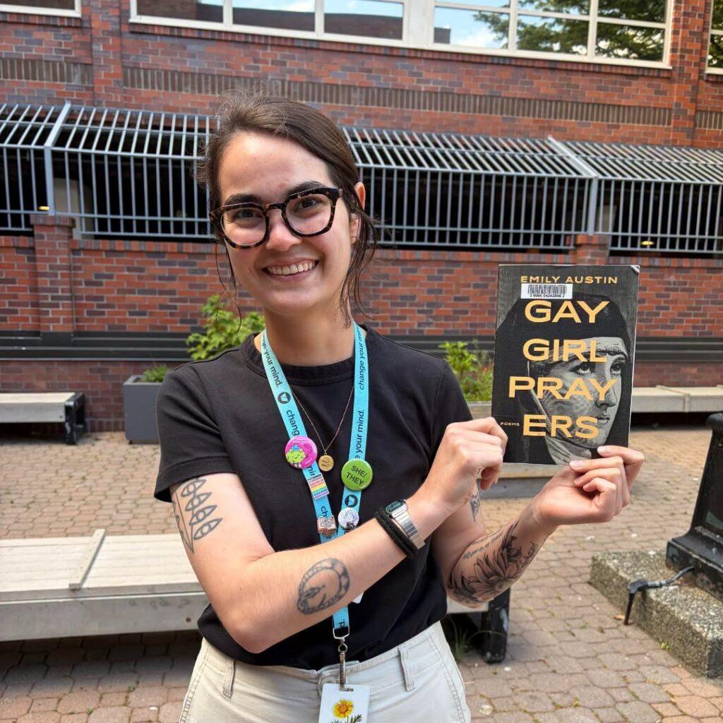 A librarian wearing glasses and a staff lanyard smiles while holding up the book “Gay Girl Prayers” by Emily Austin, standing on a brick patio outside the library.