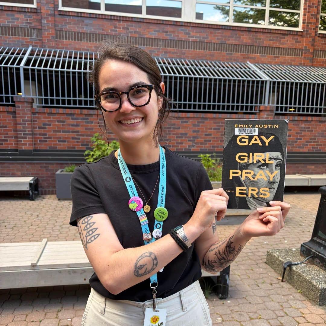 A librarian wearing glasses and a staff lanyard smiles while holding up the book “Gay Girl Prayers” by Emily Austin, standing on a brick patio outside the library.