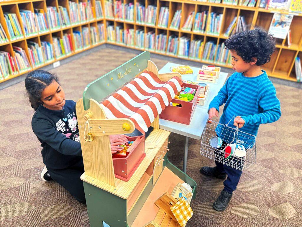 Two children playing with child-sized wooden farm market in Library. The market consists of a little stand with a wooden cash register, a table with wooden play farm goods, and a basket to put them in.