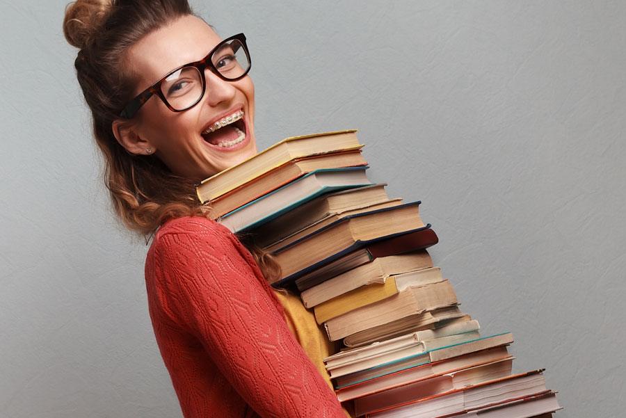 Woman with black framed glasses and a giant smile, carrying a huge pile of books.
