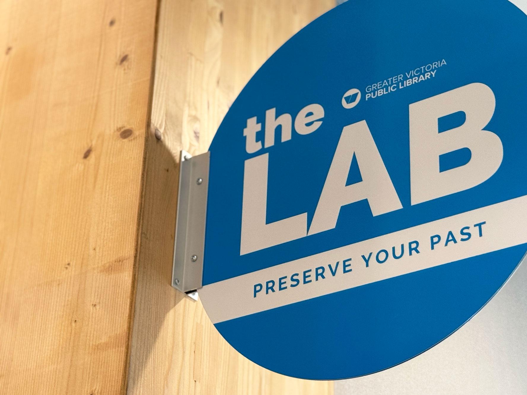 Closeup view of Esquimalt branch signage: The Lab: Preserve Your Past. A round blue panel jutting out from a cedar beam.