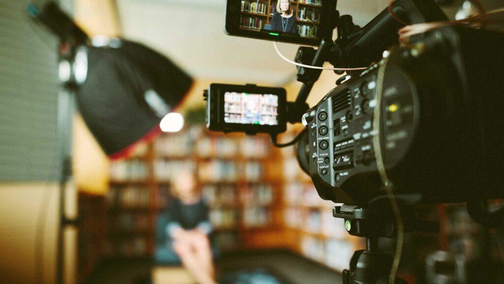 Camera in the foreground, filming a person seated in the library.
