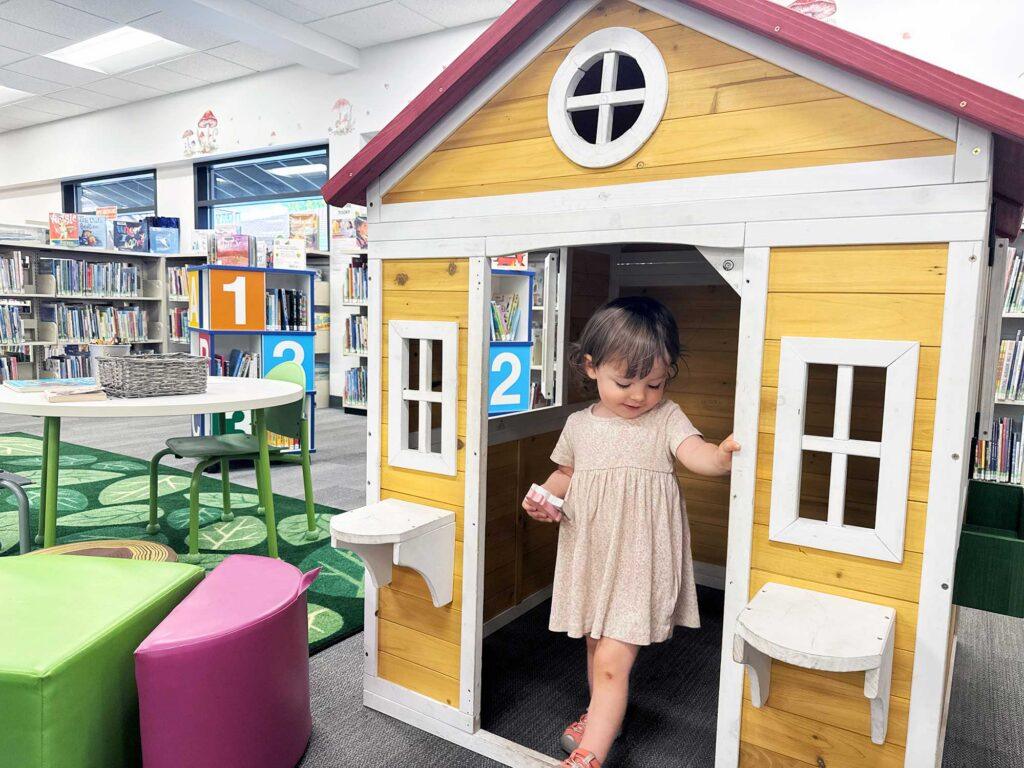 Young girl in summer dress, inside small wooden cottage playhouse in Oak Bay Library children's section. Colourful rug and furniture in the background.