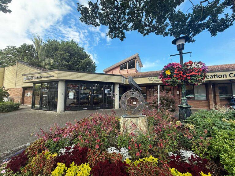Oak Bay Branch exterior with flower bed, and colourful double hanging basket in the foreground. A oak tree overhead. The building is a single storied, flat roofed structure, with cement poles.