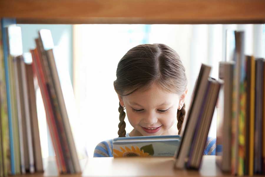 Young girl with braided hair smiling while discovering a new book, seen from through the library book shelves.