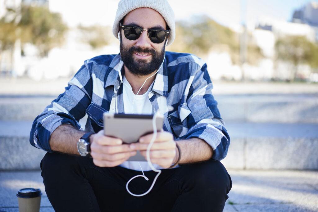 Young bearded man, sitting on sidewalk enjoying a coffee while reading his tablet.