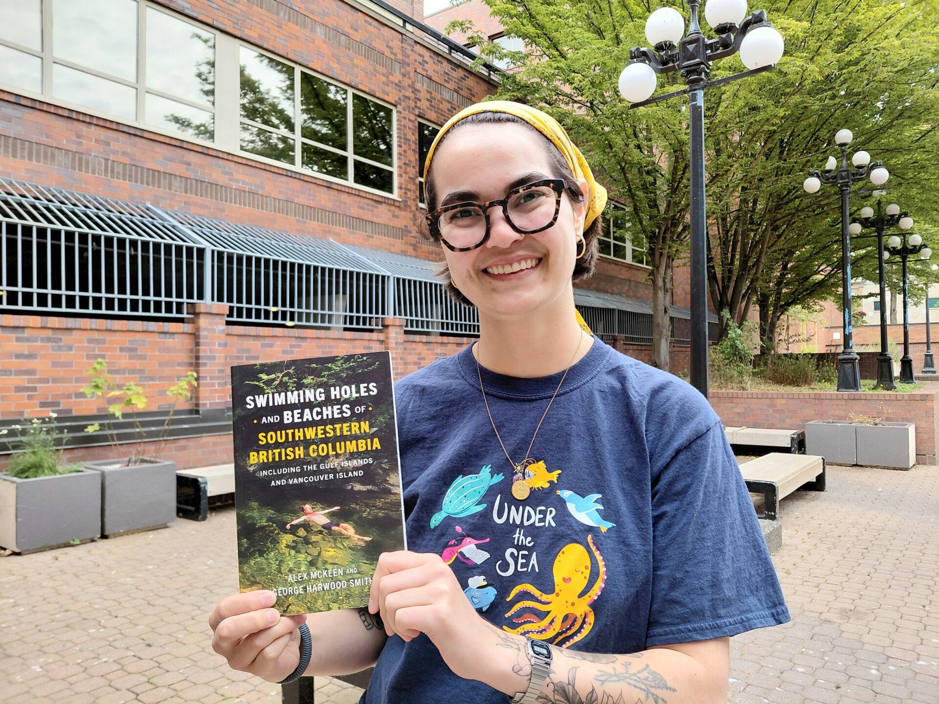 A librarian wearing glasses and summer reading club shirt holding a book.