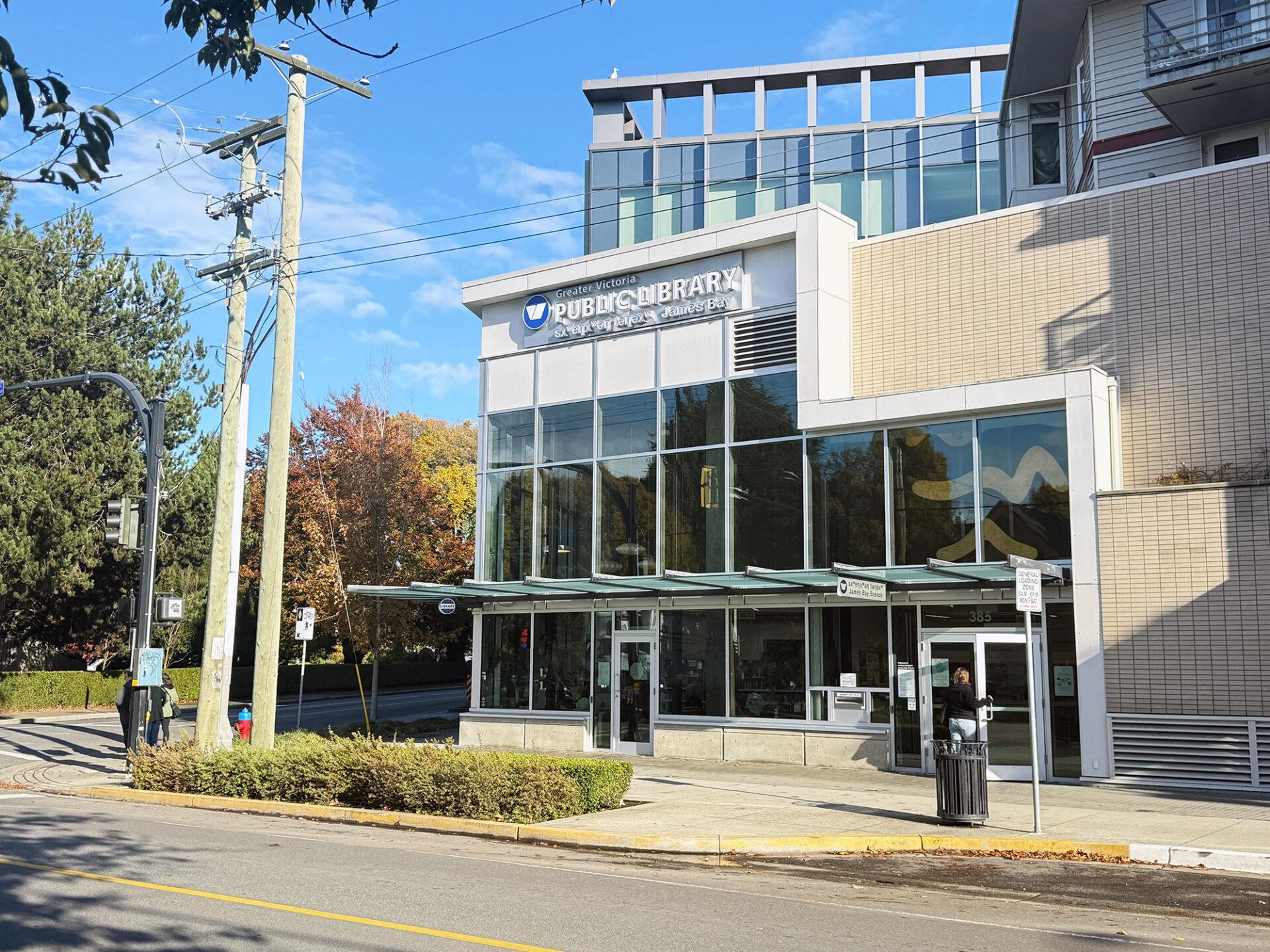 Bright sunny view of the sxʷeŋxʷəŋ təŋəxʷ James Bay Branch from the street. A corner two story building with predominantly glass windows and branch signage above.