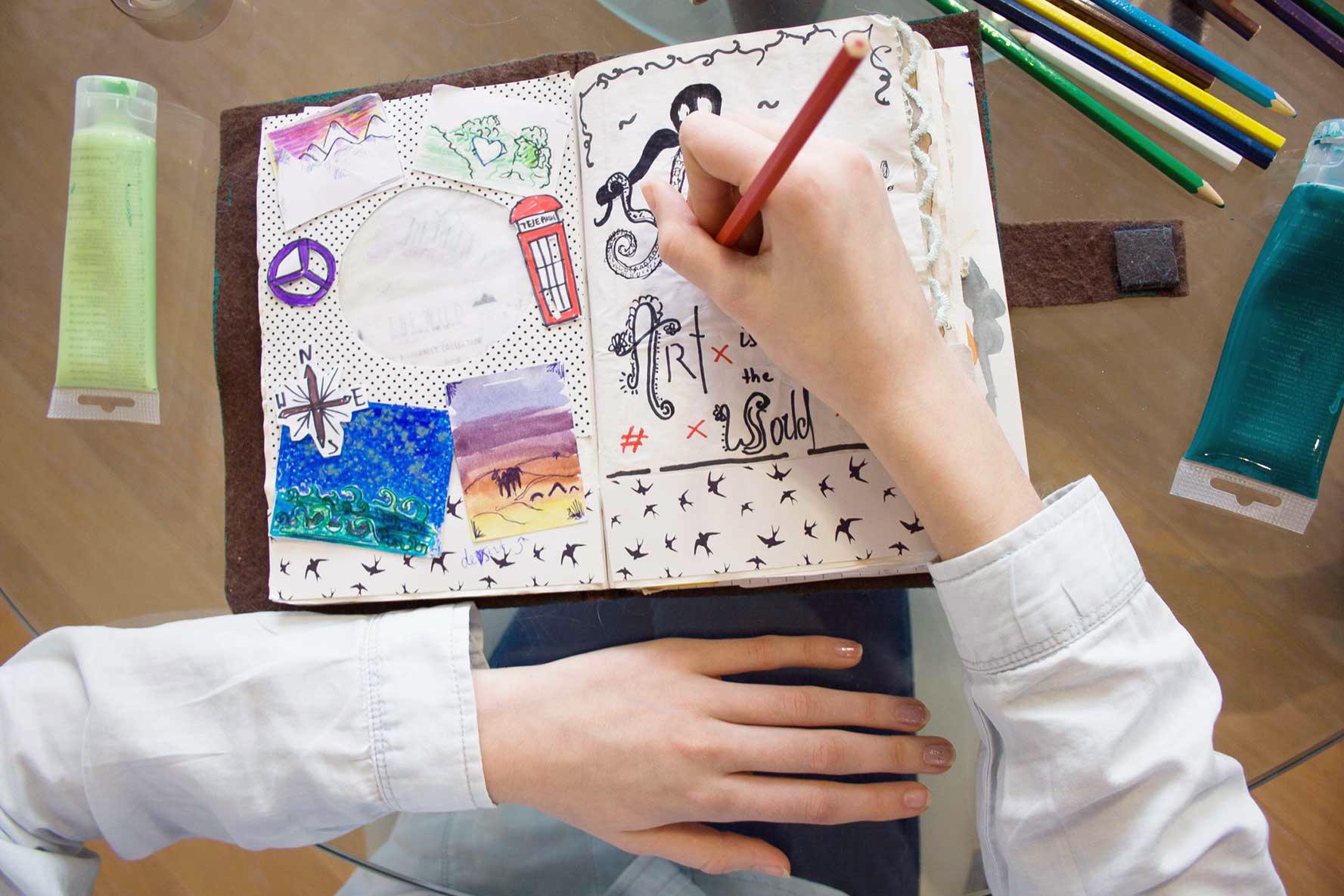 Overhead view of young hands holding a coloured pencil while drawing in a collaged journal full of stylized words and drawings.
